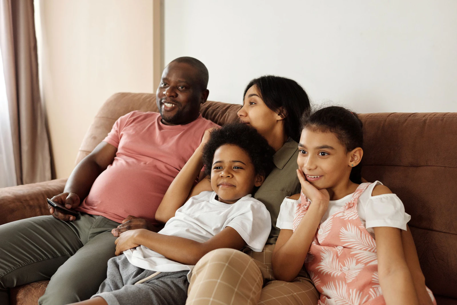 Pai, mãe e dois filhos sorrindo no sofá da sala enquanto assistem à TV, com o controle remoto na mão do pai