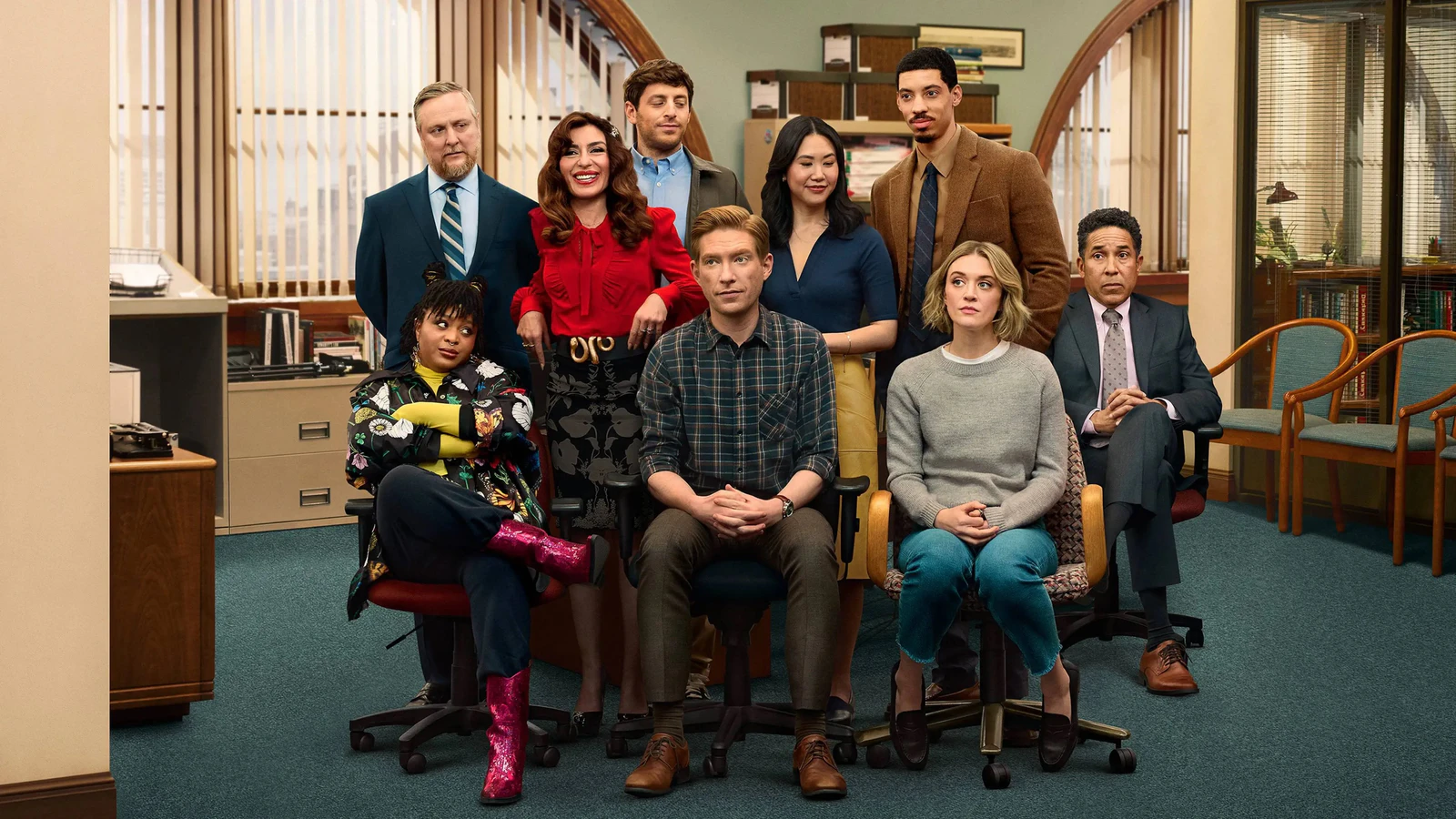 Elenco da série The Paper posando na redação do jornal, com arquivos, estantes e cadeiras de escritório ao fundo