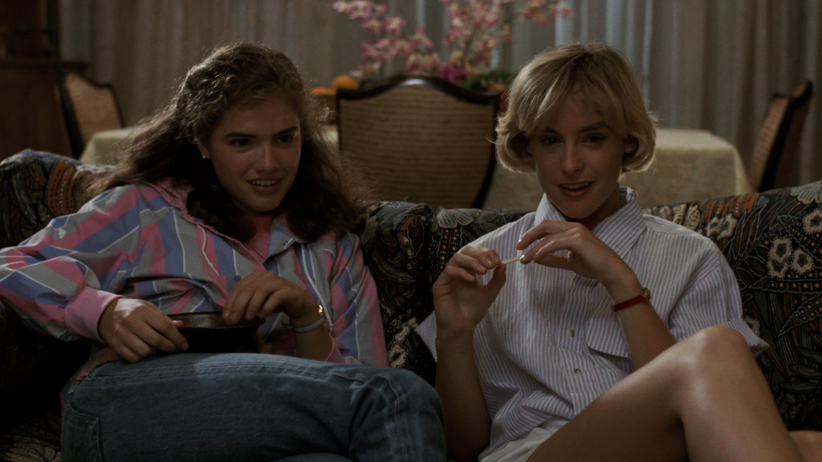 Nancy e Tina sorrindo no sofá da sala, com petiscos no colo, em cena de A Hora do Pesadelo (1984), clássico de terror na HBO Max