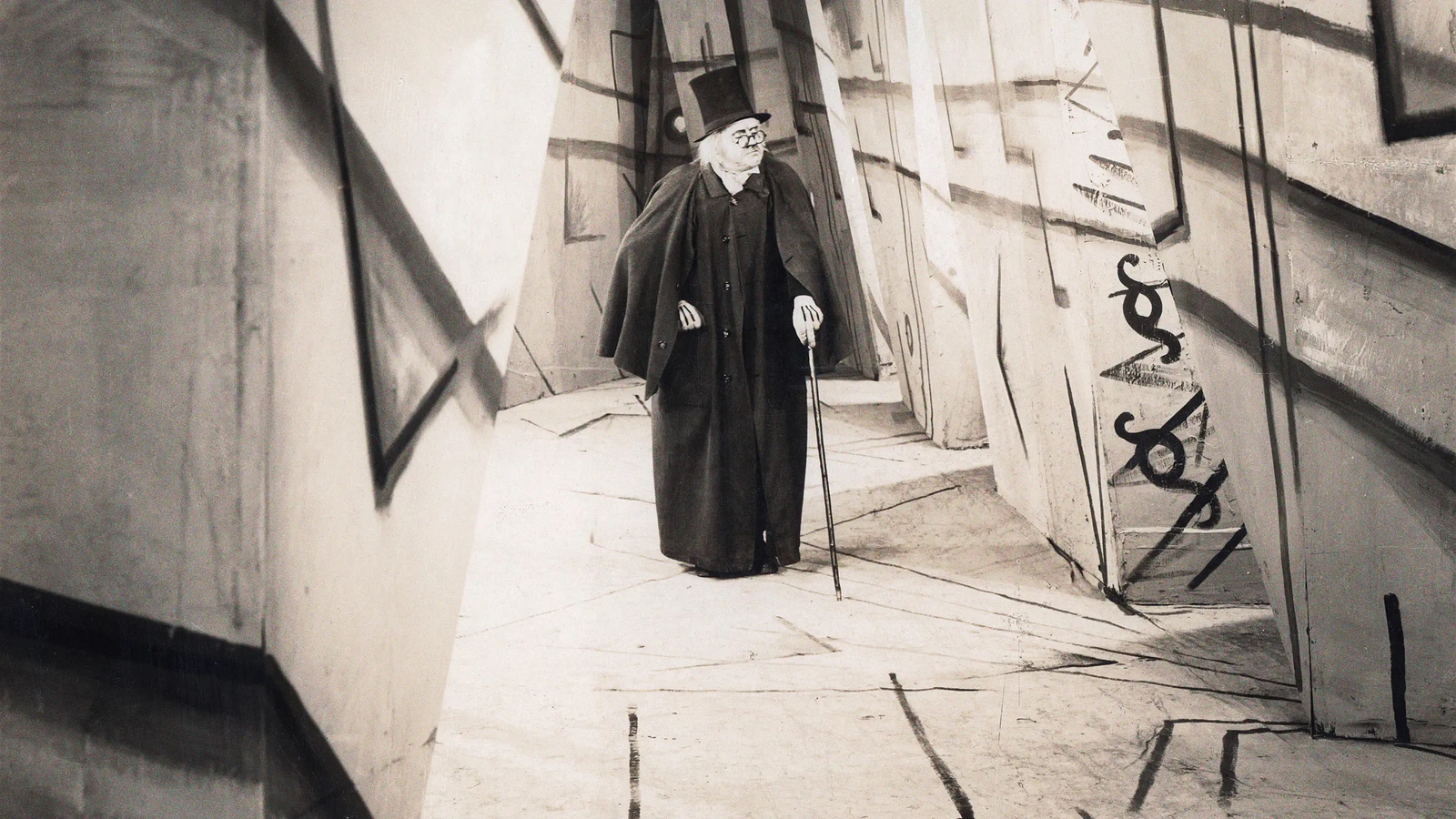 Dr. Caligari de cartola e bengala caminhando por uma viela de paredes tortas pintadas à mão, em O Gabinete do Dr. Caligari, clássico de terror no Telecine