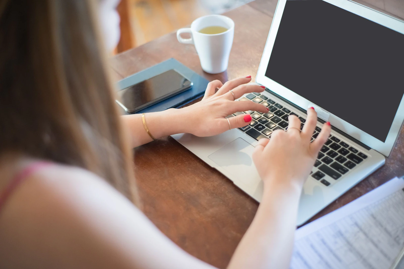 Mulher digitando no notebook numa mesa de madeira em casa, com o celular e uma xícara de chá ao lado, cenário de quem compara os planos do Gemini 4