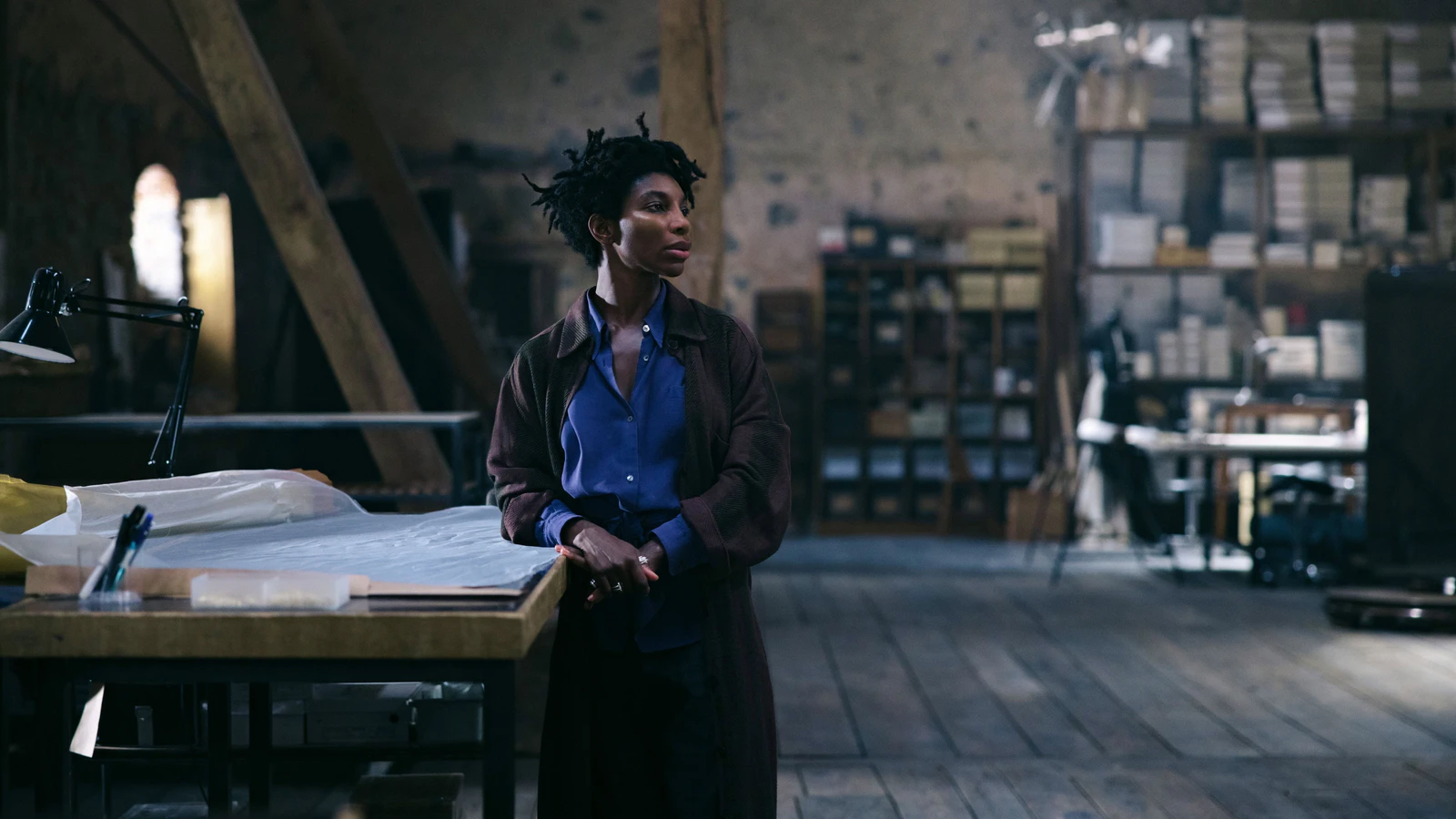 Michaela Coel de camisa azul e casaco marrom encostada numa mesa de corte num ateliê de costura com vigas de madeira e estantes de caixas, em cena de Mother Mary