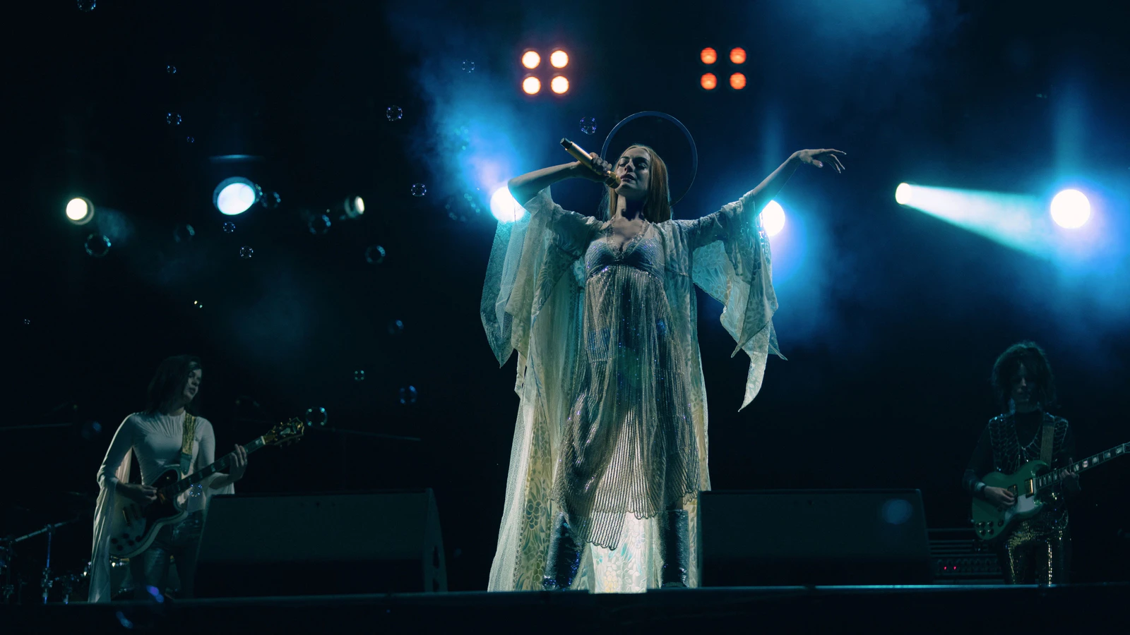 Cantora de vestido branco esvoaçante e auréola canta ao microfone no palco, com dois guitarristas e refletores azuis, em cena de show de Mother Mary